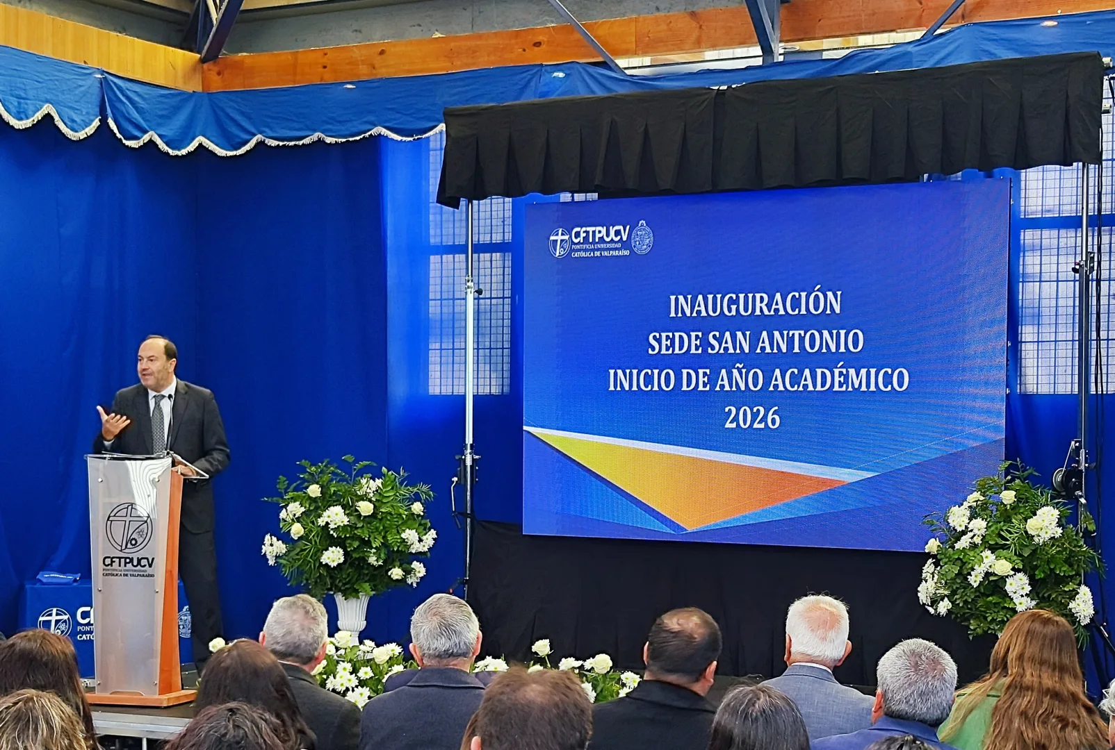 Man in a suit speaks at a clear podium during an inauguration ceremony on a blue stage backdrop.