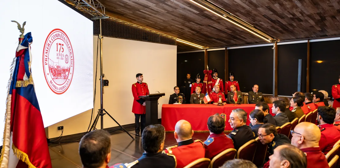 Military ceremony in a hall with uniformed personnel seated at a long red-cloth table and flags on the sides, and a large projection screen with a crest.