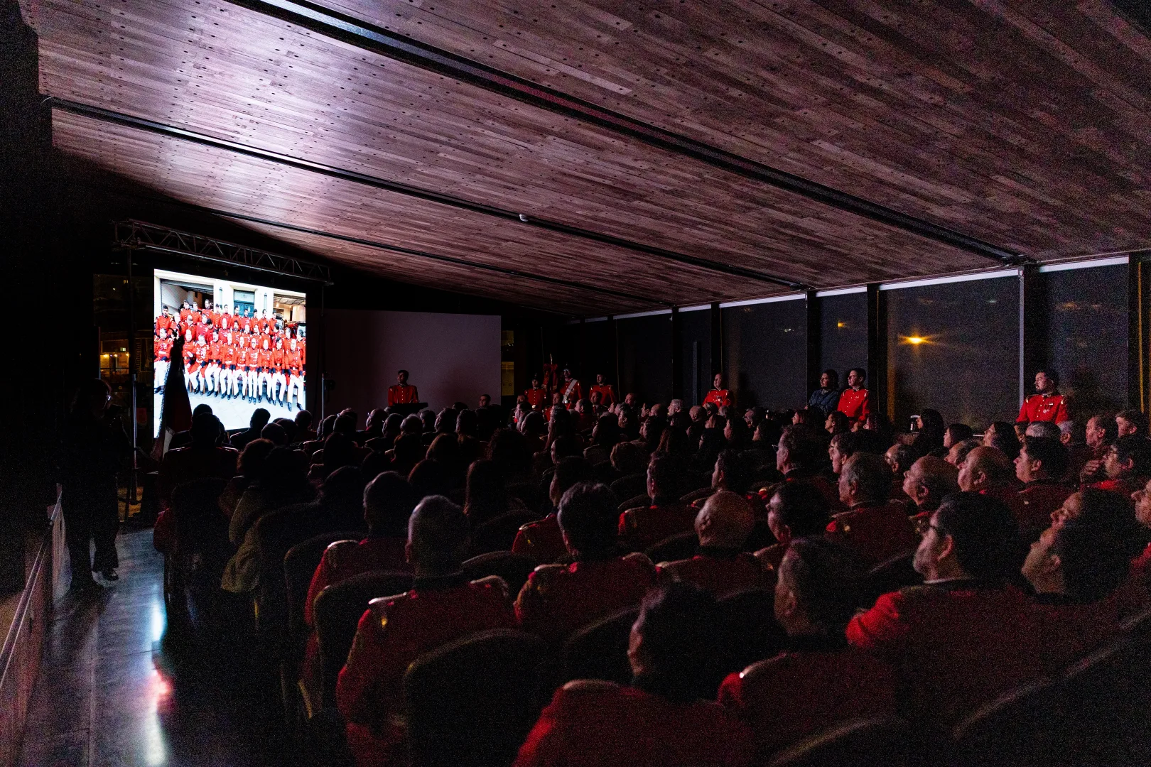 Audience seated in a dim room watching a projection of a group photo of people wearing red uniforms on a large screen to the left.