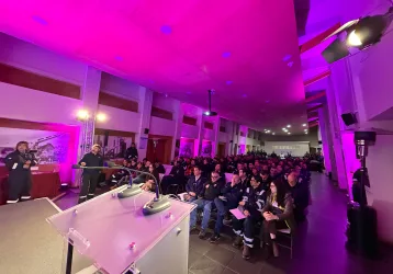 Iluminación Led para eventos corporativos - FBAudio – Pantallas Led – Streaming – Amplificación Audience seated in a large hall watching a panel at a raised stage with lectern under purple lighting