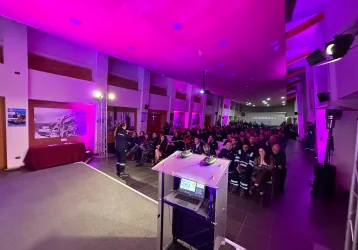 Iluminación Led para eventos corporativos - FBAudio – Pantallas Led – Streaming – Amplificación Presenter at a transparent lectern addressing a large audience in a room bathed in purple lighting.